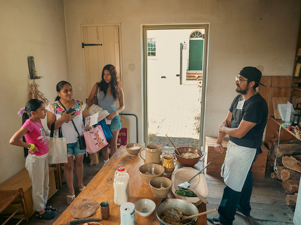 Visitors gather around a historic kitchen table as an interpreter demonstrates food preparation inside a museum's colonial-era kitchen.
