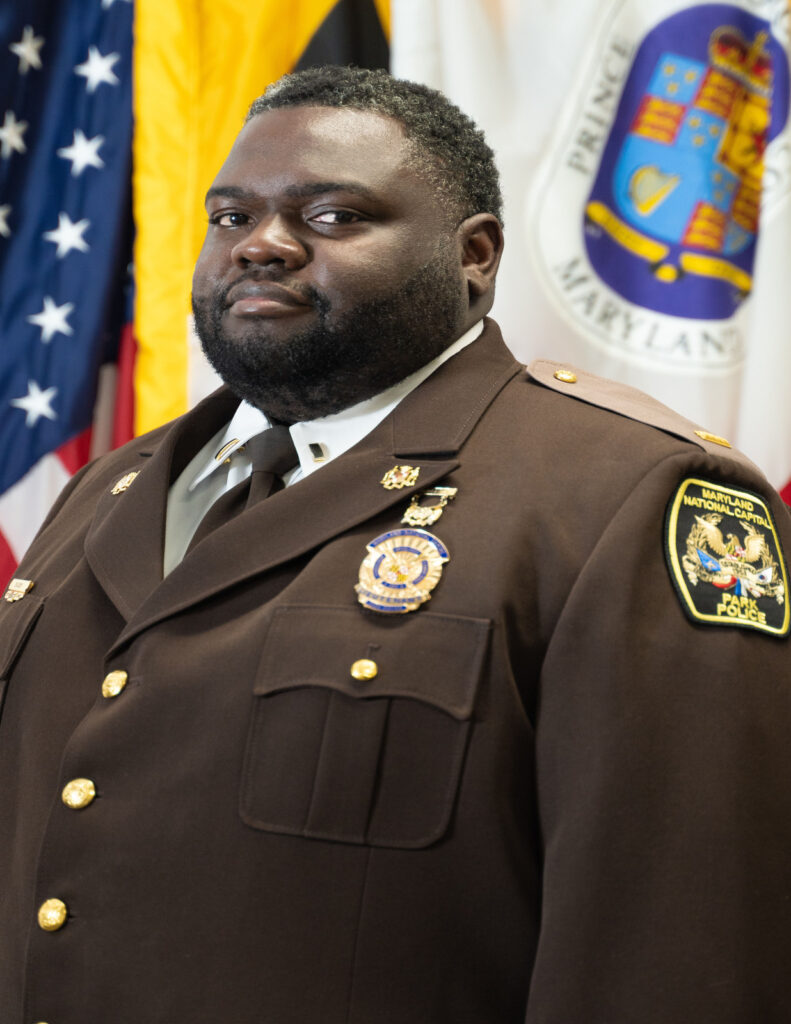 Uniformed park police official in dress uniform, photographed before American, Maryland, and Prince George's County flags.