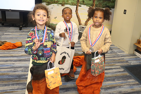 Three kids holding orange and brown bags and wearing medals around their necks. One carries a bag that says Woolly Bear Wingding.