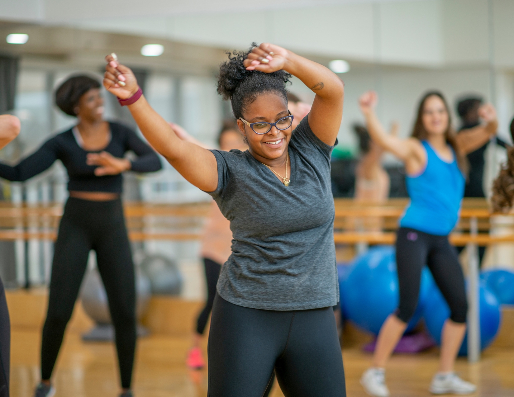 A smiling young woman dancing with arms raised during an energetic group dance exercise class in a fitness studio.
